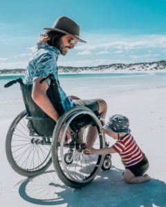 Jaimen Hudson in a Wheelchair on the beach looking down at his young toddler son playing with the wheel of his wheelchair